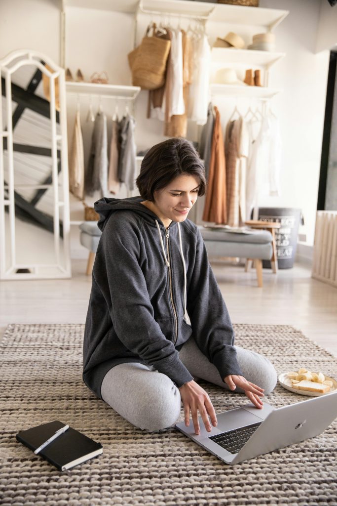 Cheerful young lady in casual wear sitting on carpet and typing on keyboard of computer while resting on floor in cozy light apartment